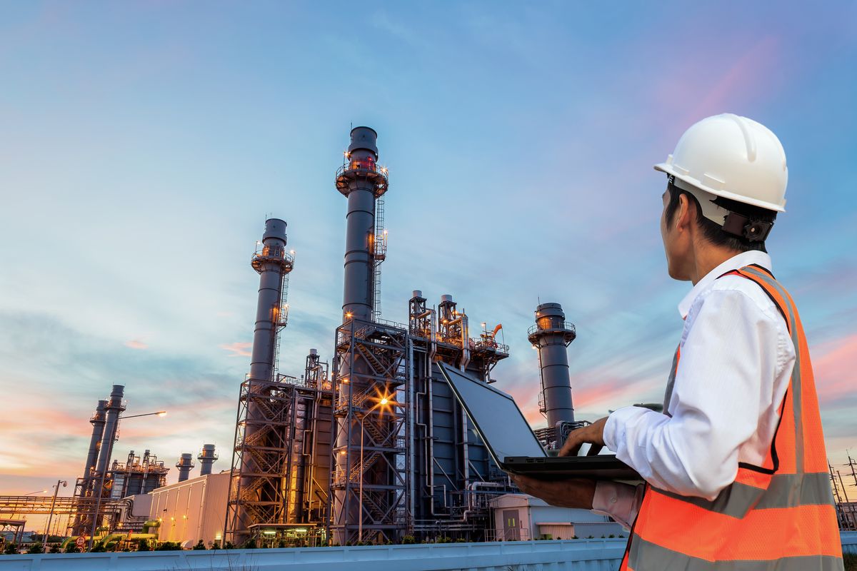 A worker in a white hard hat and orange safety vest uses a laptop while observing a large industrial facility with tall chimneys, set against a colorful sunset sky.