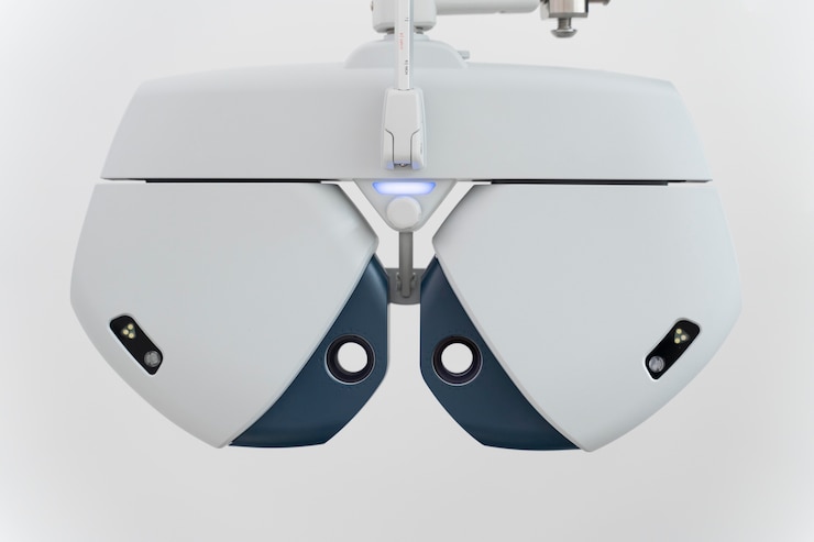A modern optometry device with a sleek design, featuring two rounded white and blue sections with small lenses and sensors, hanging from an adjustable arm against a plain background.
