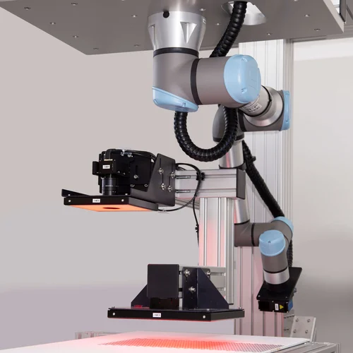 A robotic arm with blue joints is positioned above a workspace, equipped with a camera and lighting system for inspection or analysis. The setup is mounted on a metal frame, with wires neatly organized, suggesting a high-tech laboratory or industrial setting.