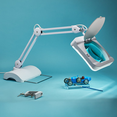 A white magnifying lamp with an adjustable arm is positioned over a small blue toy car on a light blue surface. Nearby, a small gray key and a blue tool are placed, adding to the scene's detail.