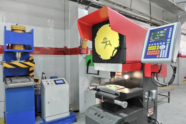 A precision measurement machine with a digital display and a yellow projection screen is set up in an industrial workshop. Next to it is a blue hydraulic press with a control panel, surrounded by a clean, organized workspace with a red and white wall.