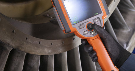 A person wearing black gloves holds an orange handheld device with a screen and buttons, inspecting a large industrial turbine with metal blades.