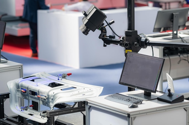 A high-tech manufacturing setup features a metal automotive component on a workstation, surrounded by computer monitors and a camera mounted on an adjustable arm, indicating a precision inspection or analysis process.