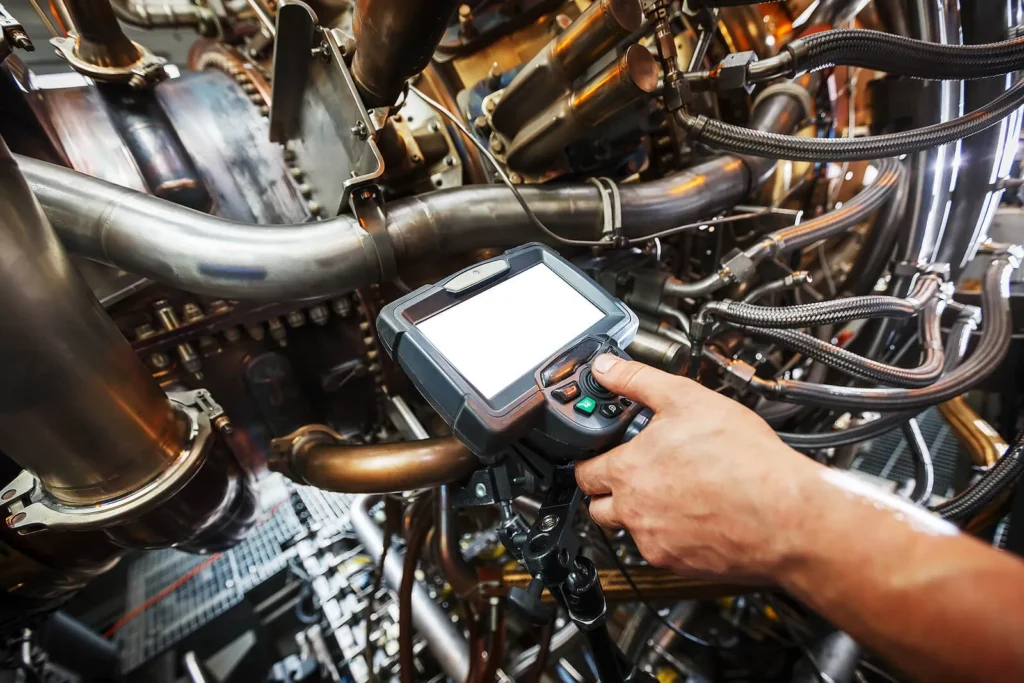 A person operates a handheld diagnostic device amidst a complex network of metal pipes and machinery, focusing on the screen while surrounded by intricate industrial components.
