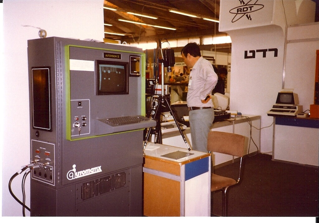 A man stands at a trade show booth featuring vintage computer equipment. On the left, a large Automonix machine with a screen and keyboard is visible.