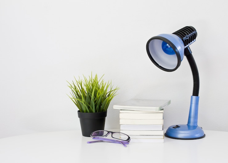 A blue desk lamp with a flexible neck is positioned beside a stack of books on a white surface. Nearby, a small black pot holds a vibrant green plant, and a pair of glasses rests in front of the books.