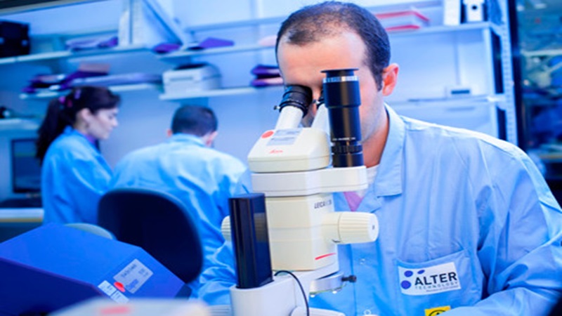 A scientist in a blue lab coat is intently looking through a microscope in a laboratory setting, with two colleagues working in the background. Shelves with equipment and documents surround them, creating a busy research environment.