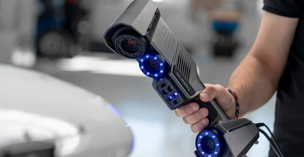 A person holds a handheld 3D scanner with multiple lenses and glowing blue lights, set in a modern, industrial environment.