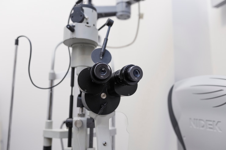 A close-up view of an ophthalmic slit lamp with two adjustable eyepieces, set in a clinical environment with a Nidek machine partially visible in the background.