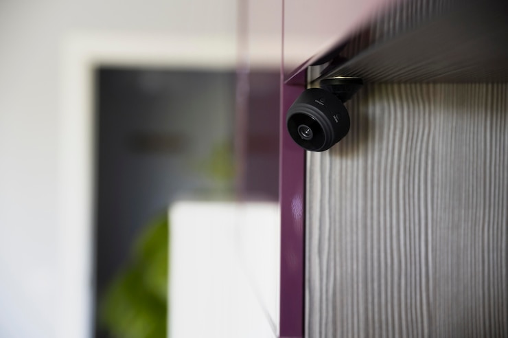 A small black security camera is mounted on the corner of a glossy purple cabinet, set against a wood-textured wall.