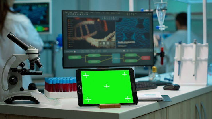 A laboratory scene featuring a microscope, a tablet with a green screen, and test tubes filled with red liquid on a countertop, with a computer displaying DNA analysis in the background.