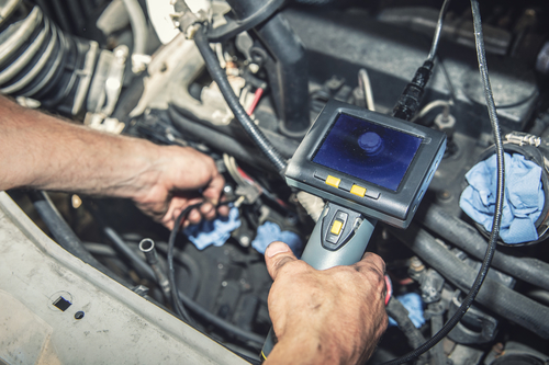 A person uses a handheld diagnostic tool with a screen to inspect the engine of a vehicle, surrounded by various cables and components, with a blue cloth nearby.