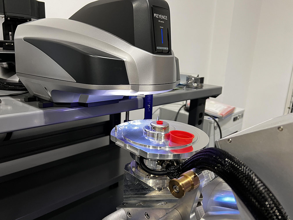 A Keyence measuring instrument is positioned over a circular metal platform with a red component, illuminated by blue light. The setting is a laboratory environment with various equipment visible in the background.