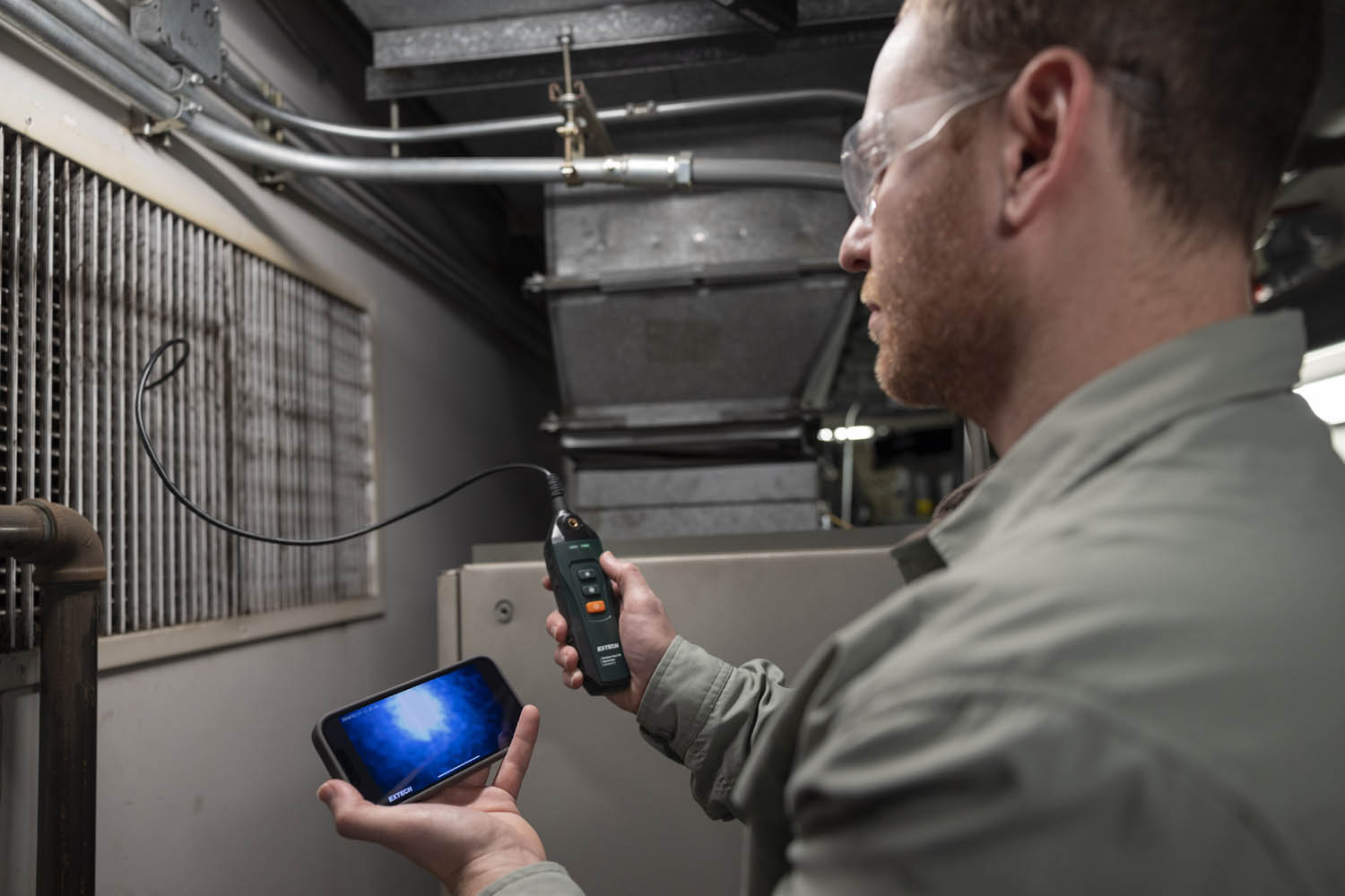 A person wearing safety goggles uses a handheld inspection camera with a flexible probe, viewing its display on a smartphone. The setting appears to be an industrial or maintenance area with metal pipes and a large vent.