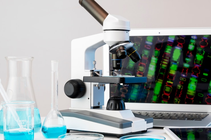 A microscope is positioned on a lab table alongside glassware containing blue liquid, with a laptop displaying colorful scientific images in the background.