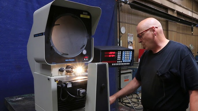 A man operates a Deltronic machine with a digital display in an industrial setting, wearing glasses and a black shirt.