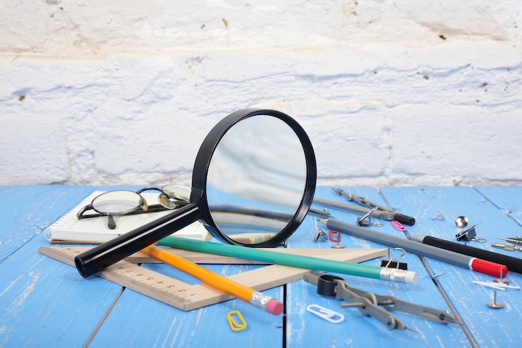 A magnifying glass with a black handle lies on a blue wooden surface surrounded by various stationery items, including pencils, a ruler, paper clips, a pair of glasses, and a compass, against a white brick wall backdrop.