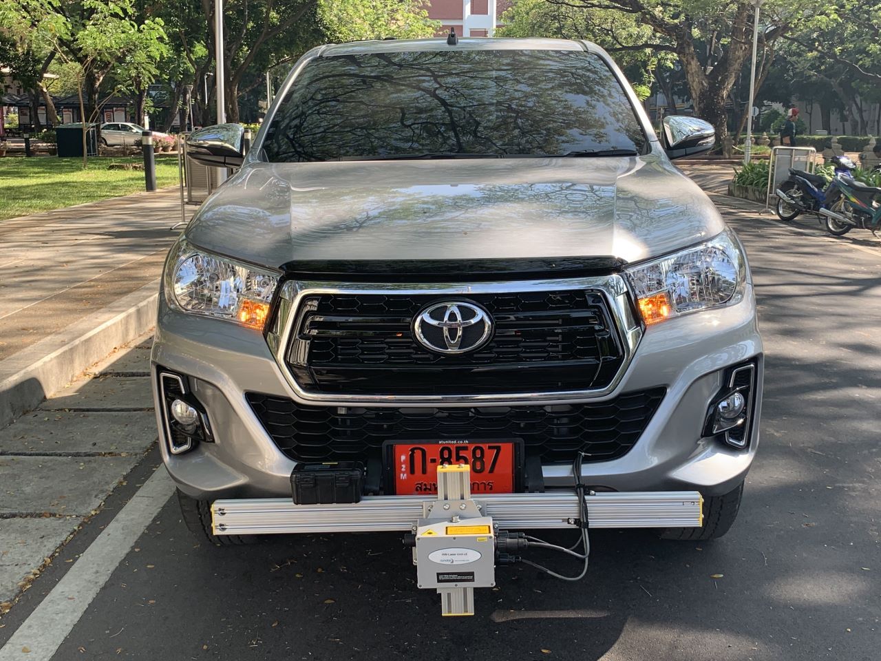 A silver Toyota pickup truck is parked on a street, equipped with a device attached to its front bumper. The vehicle has a red license plate, and the scene is set in a leafy area with trees and a sidewalk visible in the background.