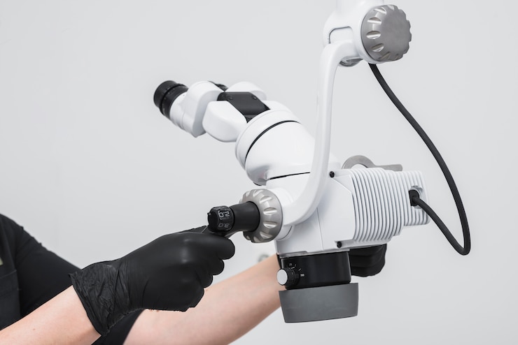 A person wearing black gloves adjusts a white and black dental microscope with multiple lenses and a flexible arm, set against a plain gray background.